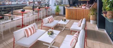 Terraza en la azotea con sofás blancos, sillas rojas y mesas, con un bar llamado "LA CERVESERIA" y vistas a la ciudad.