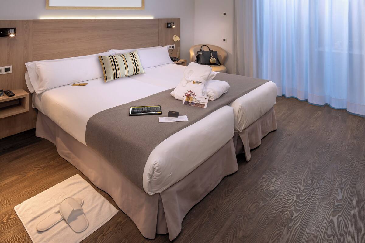 A neatly made bed with a remote control, a book, and a pair of slippers on a mat.