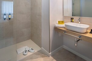 A modern bathroom with a beige tiled floor, a white sink with a wooden countertop, a glass shower enclosure, and a towel rack with two bottles.