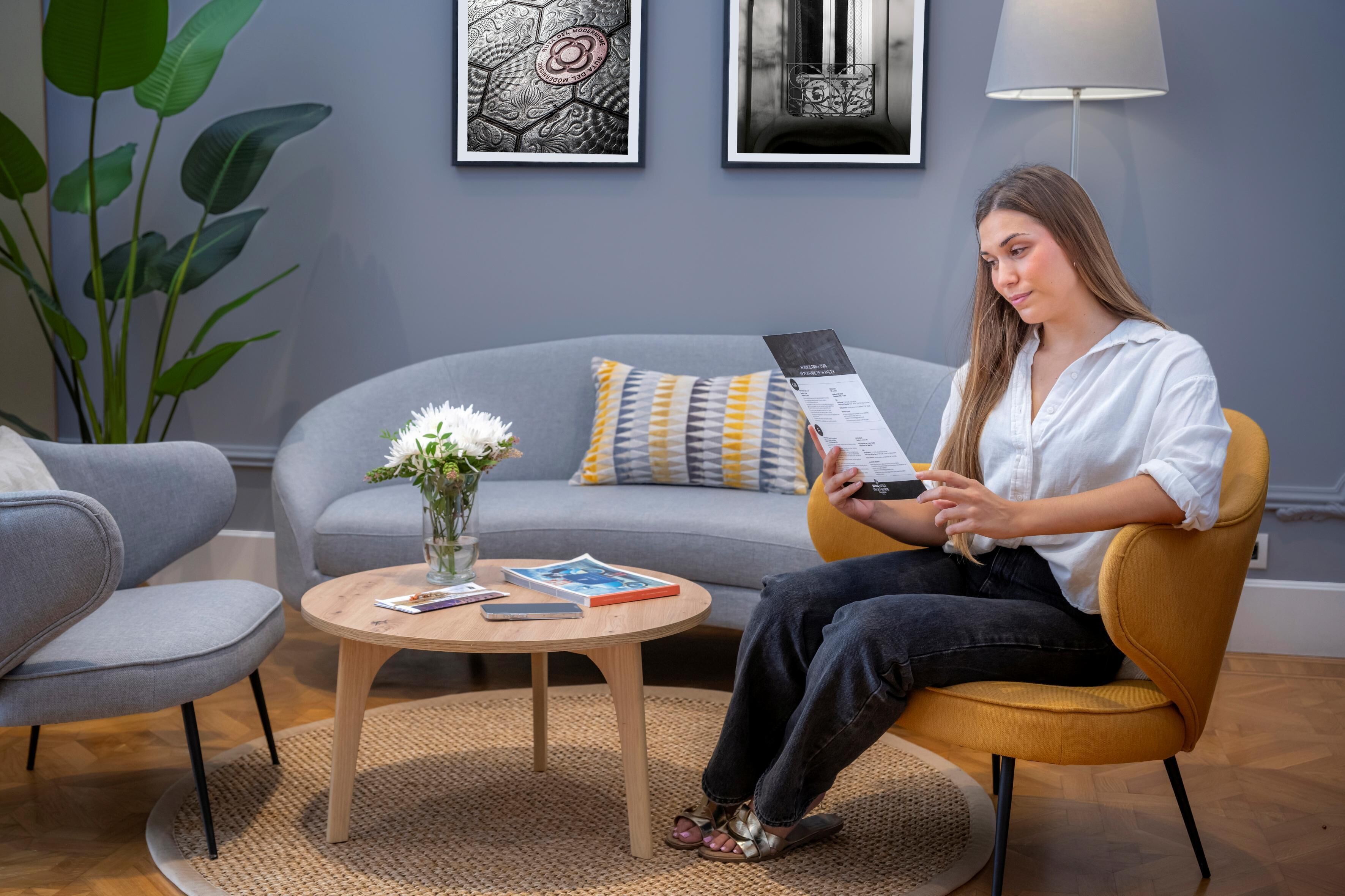 A woman sitting on a yellow chair in a modern living room with a grey sofa, a round wooden coffee table, and a vase with white flowers.