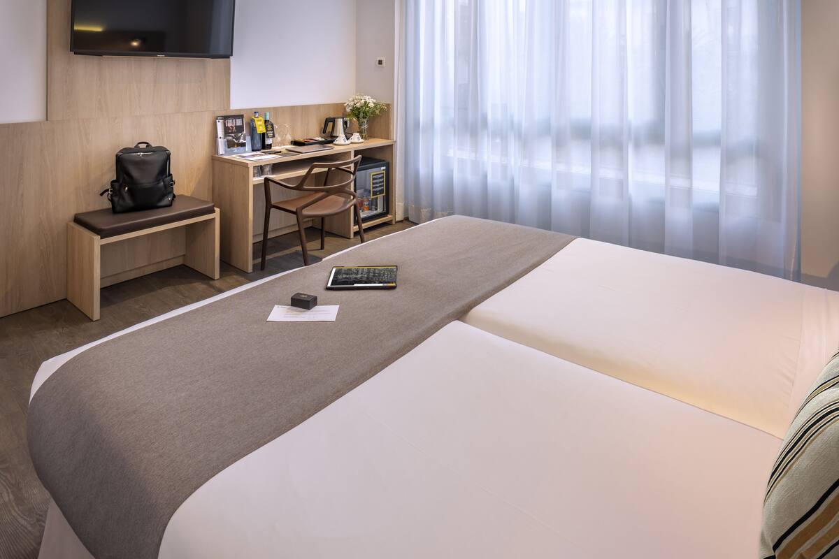 A hotel room with a bed, a desk with a chair, a small table, a TV, and a window with sheer curtains.