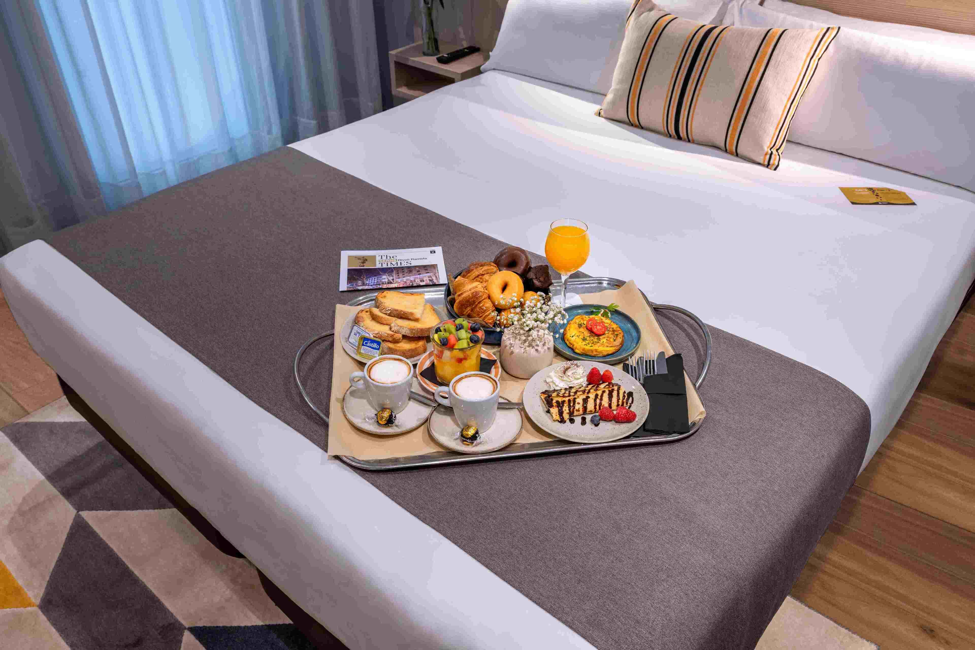 Ein ordentlich gedecktes Frühstücksbrettchen auf einem Bett mit Croissants, einer Obstschale, einem Glas Orangensaft und einer Platte mit Dessert.