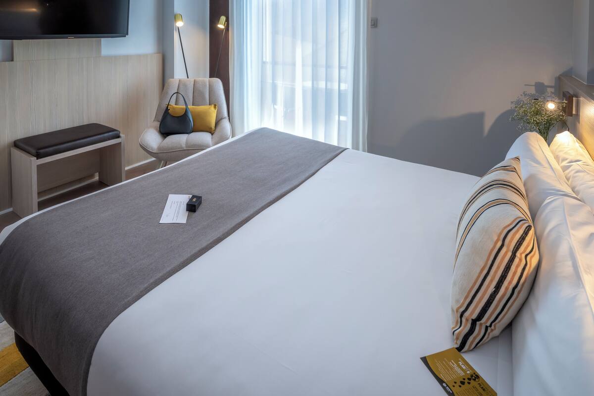 A modern hotel room with a large bed, a bedside table, a chair, a flat-screen TV mounted on the wall, and a window with sheer curtains.