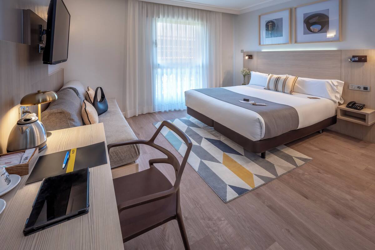 A modern hotel room with a large bed, a desk with a laptop, a chair, a coffee table, and a TV.