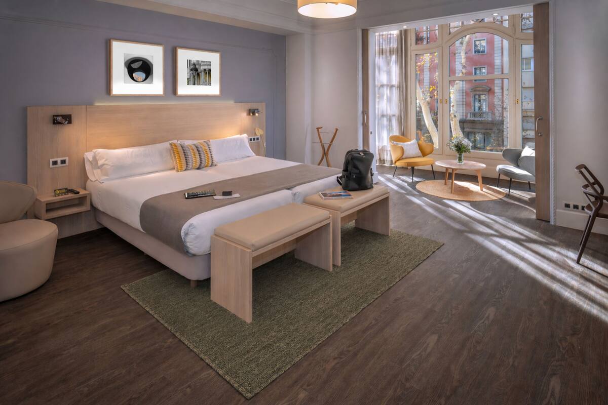 A modern hotel room with a large bed, bedside tables, a bench, a small table, and a view of the city through large windows.