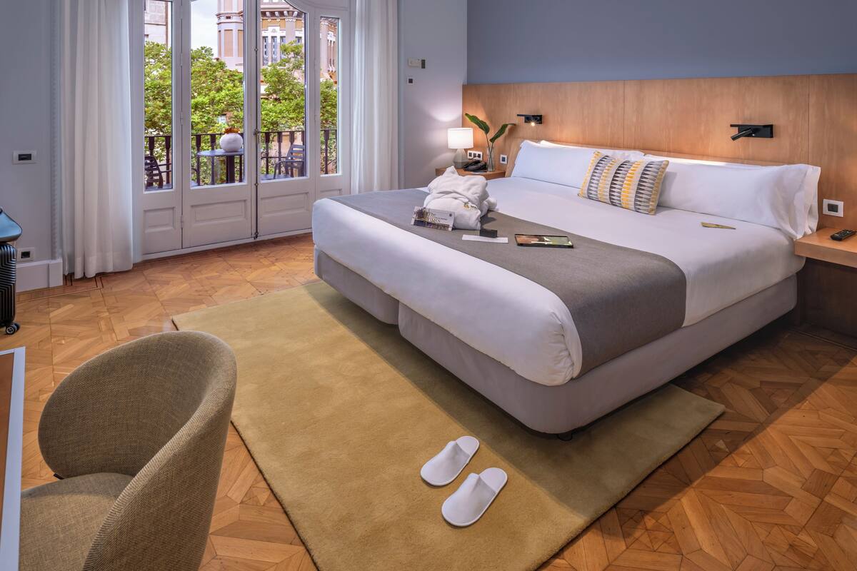 A hotel room with a large bed, a wooden headboard, a nightstand with a lamp, and a view of the outdoors through a sliding door.
