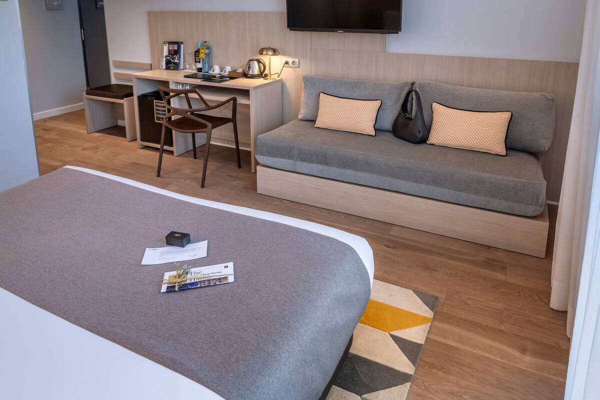 A hotel room with a large bed, a sofa, a desk with a chair, a television, and a small kitchen area.