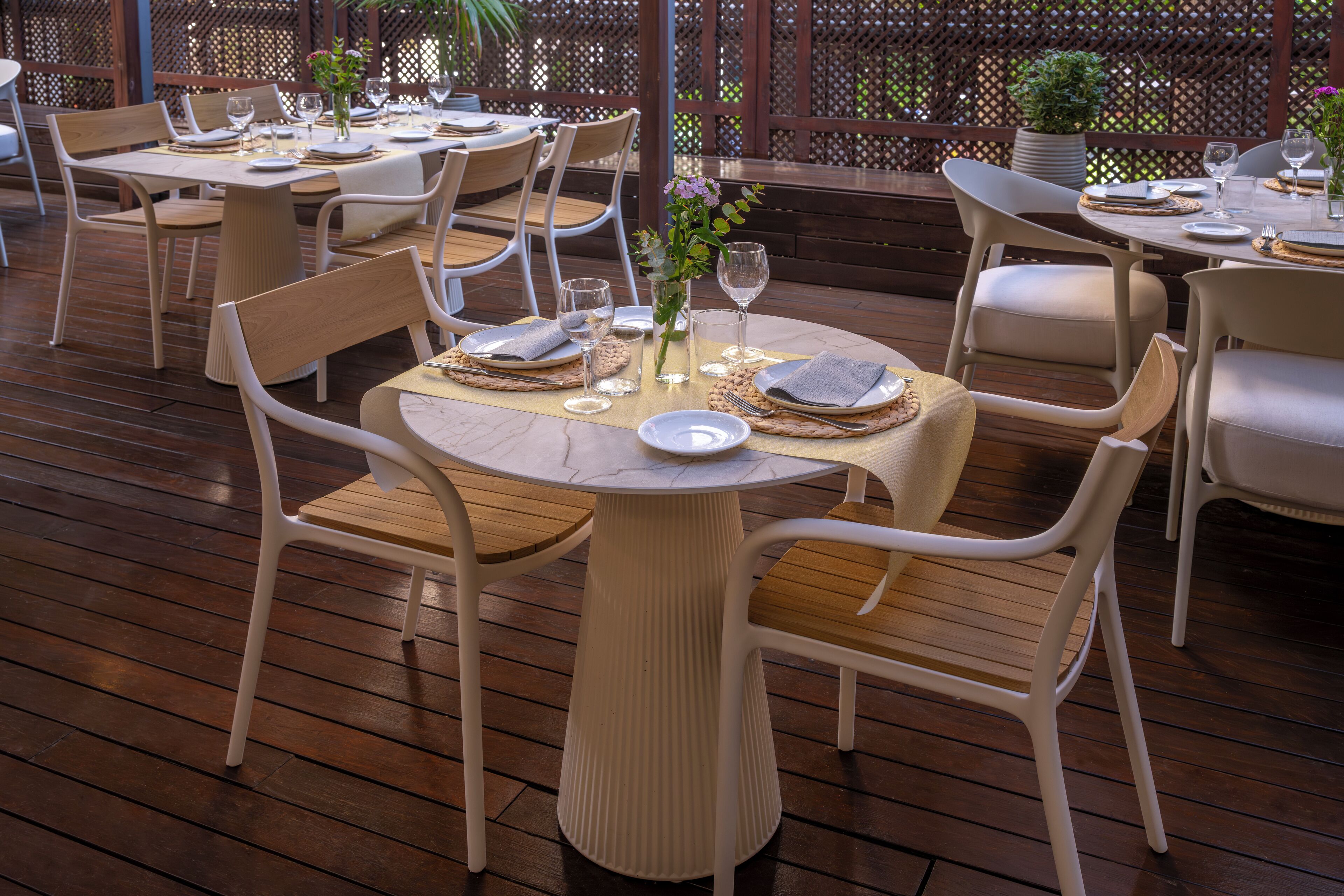 Un área de comedor al aire libre bien acondicionada con mesas de madera y sillas blancas, rodeada por una terraza de madera y un cerramiento de celosía.