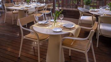 Un área de comedor al aire libre bien acondicionada con mesas de madera y sillas blancas, rodeada por una terraza de madera y un cerramiento de celosía.