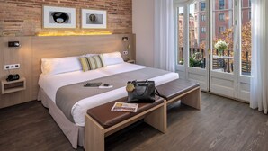 Una habitación de hotel moderna con una pared de ladrillo, una cama grande con cabecera de madera, un banco con una revista y una ventana con vistas a edificios y árboles.