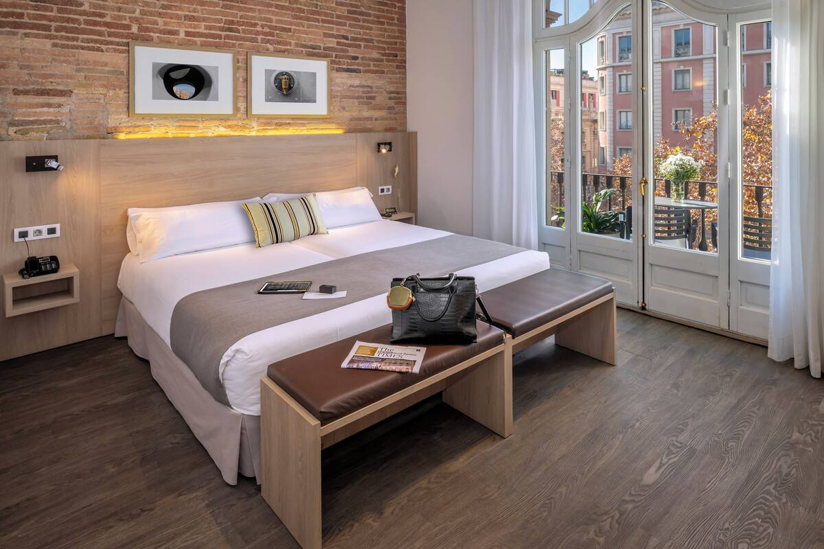 A modern hotel room with a brick wall, a large bed with a wooden headboard, a bench with a magazine, and a window with a view of buildings and trees.