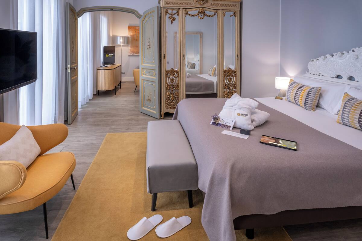 A hotel room with a large bed, a TV, a wardrobe, and a seating area.
