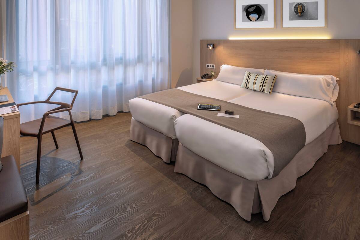A hotel room with a large bed, a desk, a chair, and a window with sheer curtains.