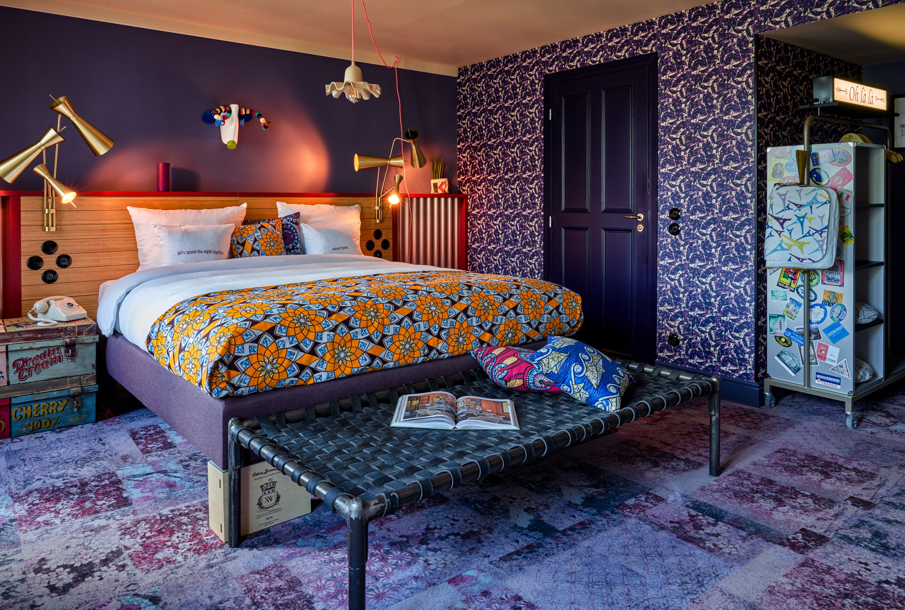 A modern bedroom with a bed, bedside lamps, a bench, and a door with a patterned wallpaper.
