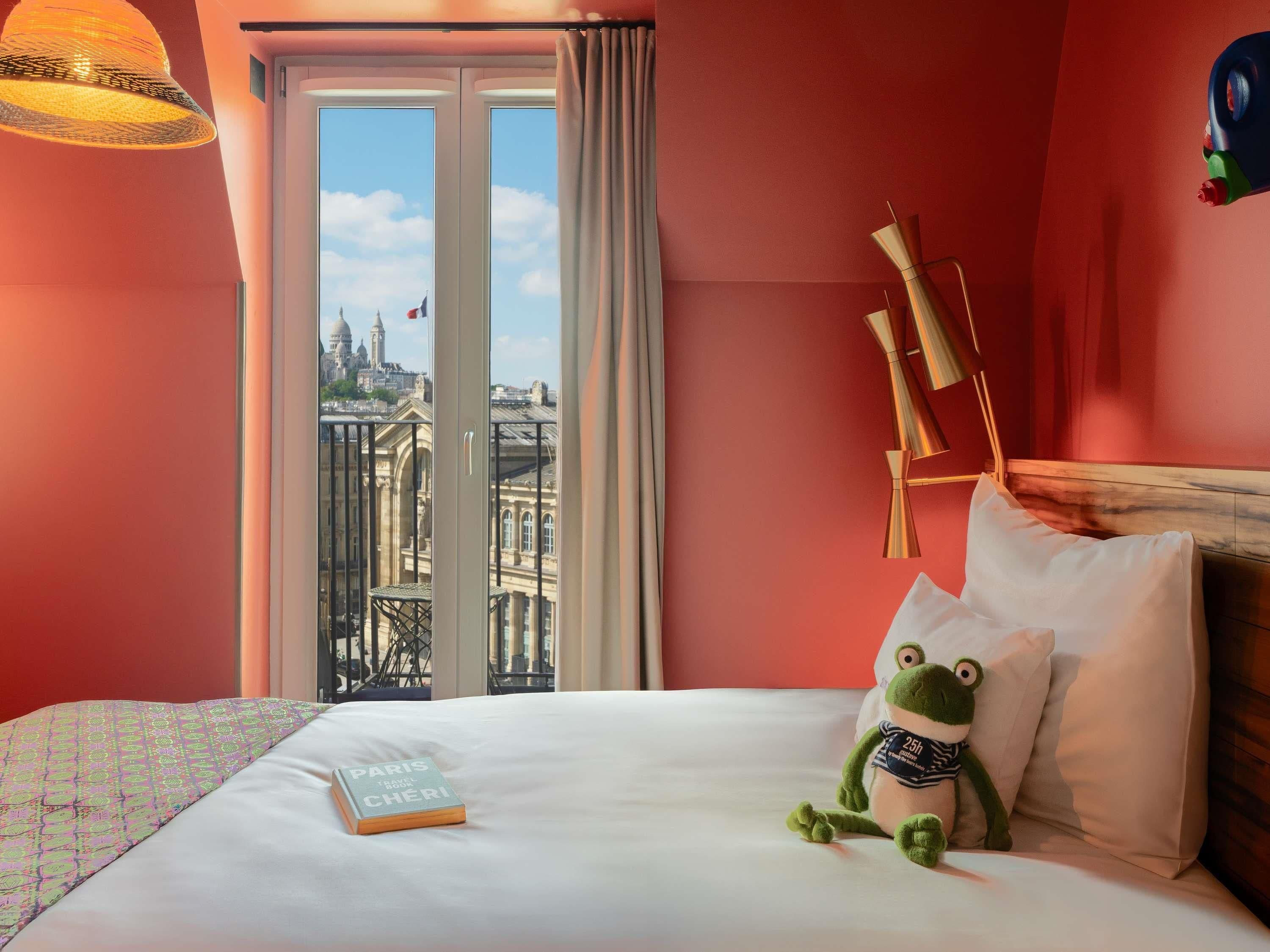 A hotel room with a bed, a plush frog toy, a book titled 'PARIS TRAVEL BOOK CHERI', and a view of a cityscape through a large window.