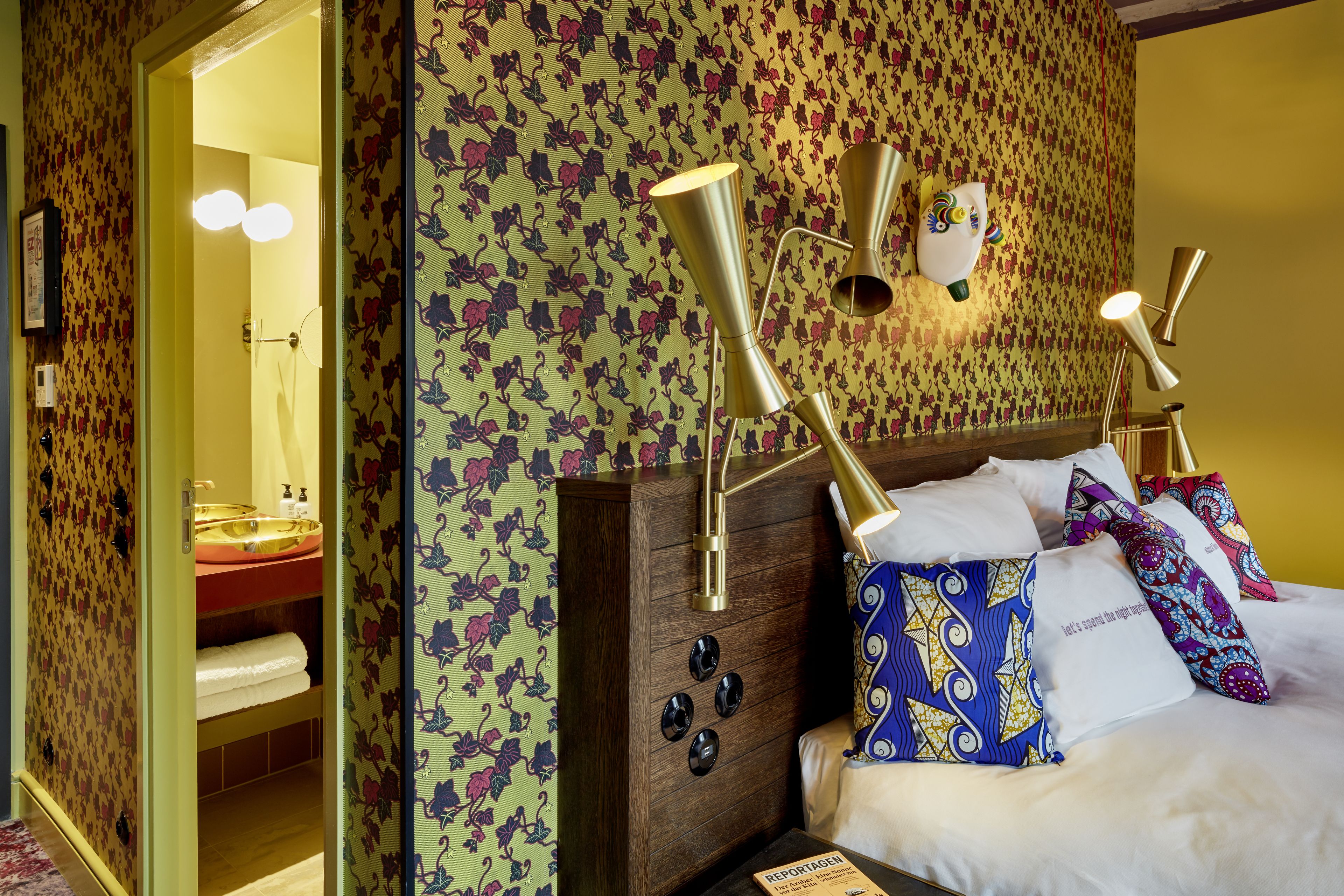 A hotel room with a wooden headboard, patterned wallpaper, and a bed with multiple pillows. There are wall-mounted lamps and a visible bathroom with a sink and towel rack.