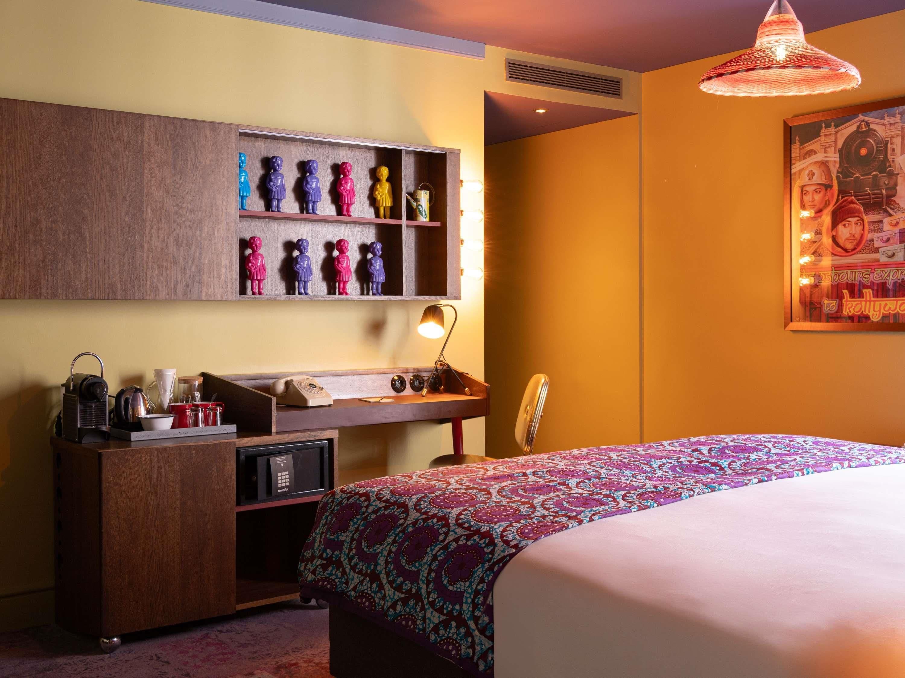 A hotel room with a bed, a desk with a lamp, a shelf with figurines, and a wall-mounted picture.