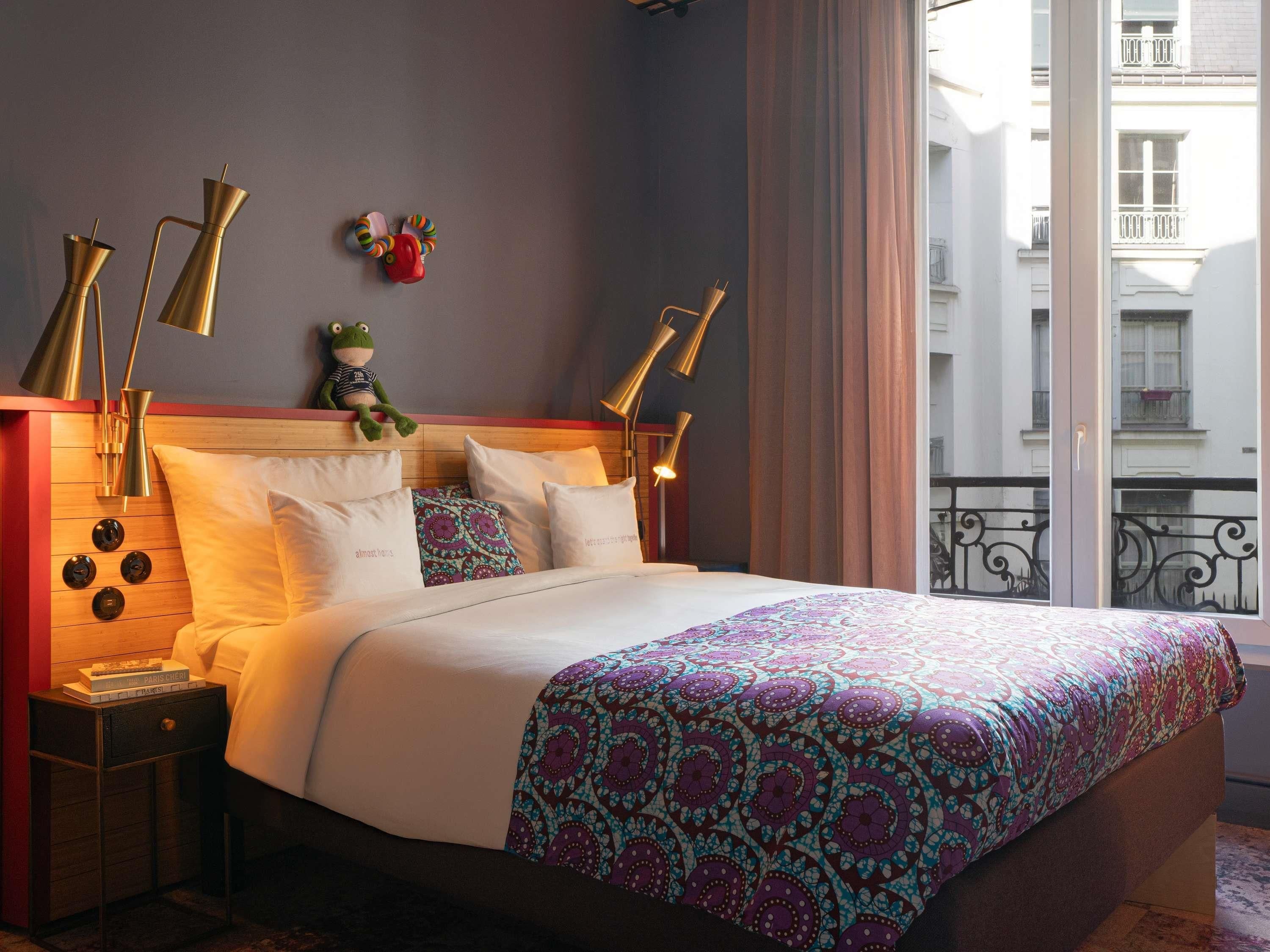 A hotel room with a bed, bedside lamps, a nightstand, a window with a view of buildings, and a balcony railing.