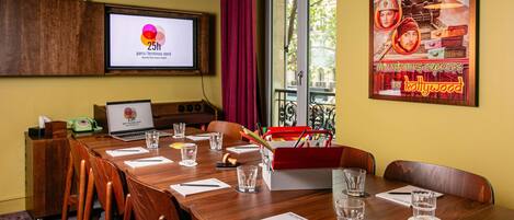 A conference room with a wooden table, chairs, a laptop, a projector screen displaying a hotel name, and a poster on the wall.