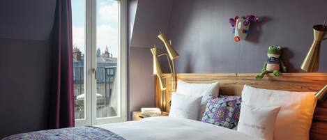 A hotel room with a large bed, a view of the city through the window, a decorative hanging light, and a small frog figurine on the headboard.