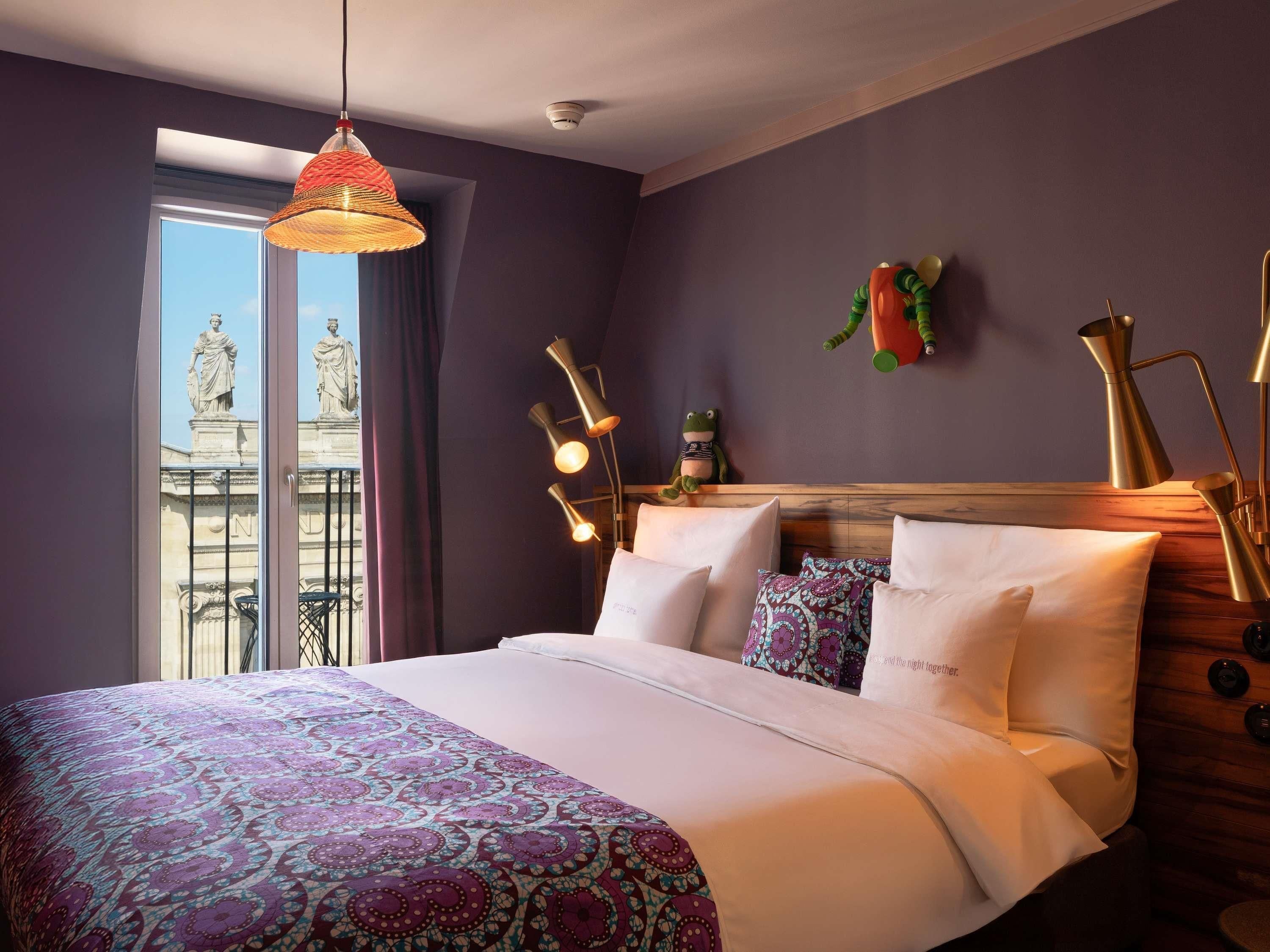 A hotel room with a bed, a window with a view of statues, a wall-mounted lamp, and a decorative frog.