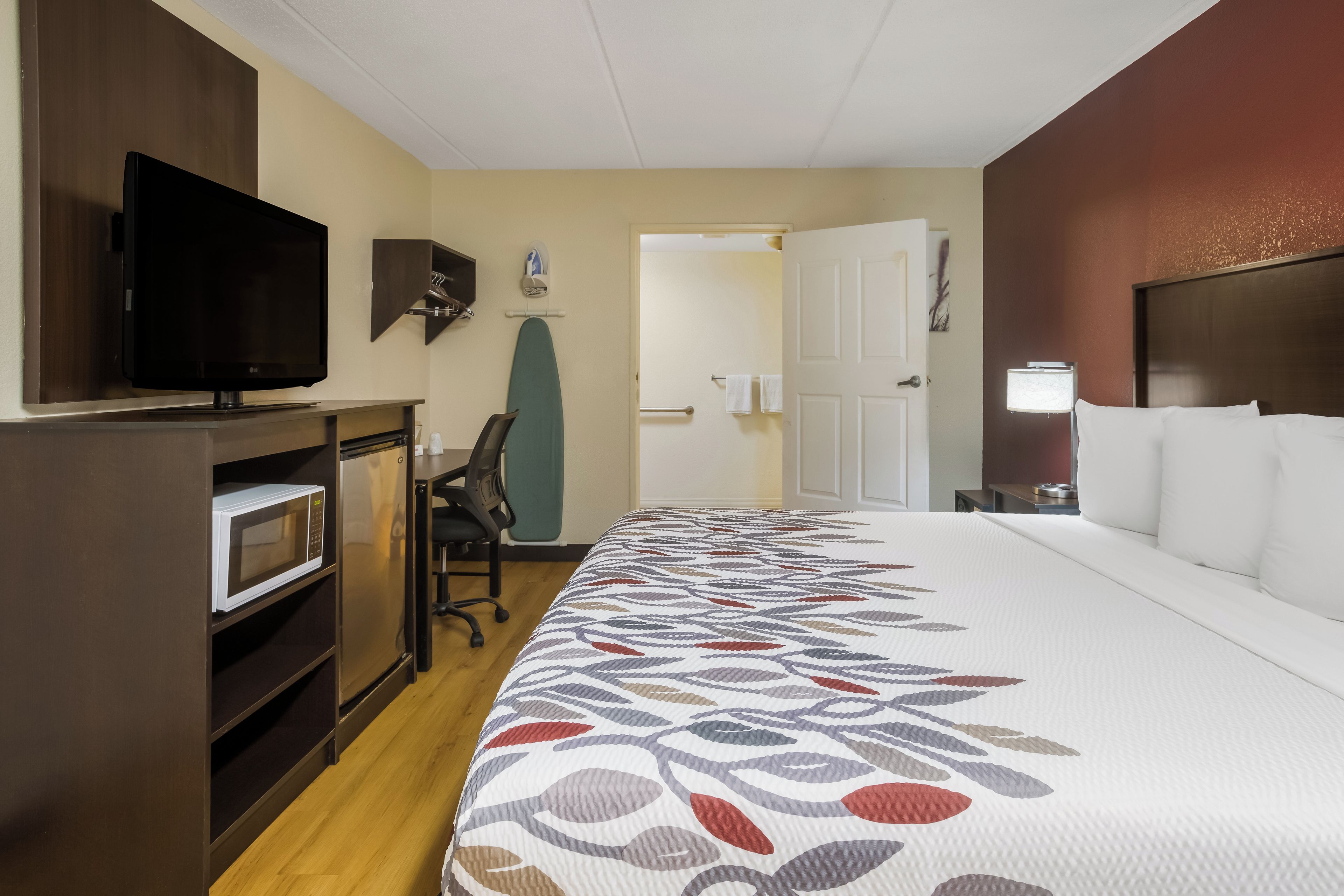 A hotel room with a bed, a television, a microwave, a refrigerator, an ironing board, and a door leading to another room.