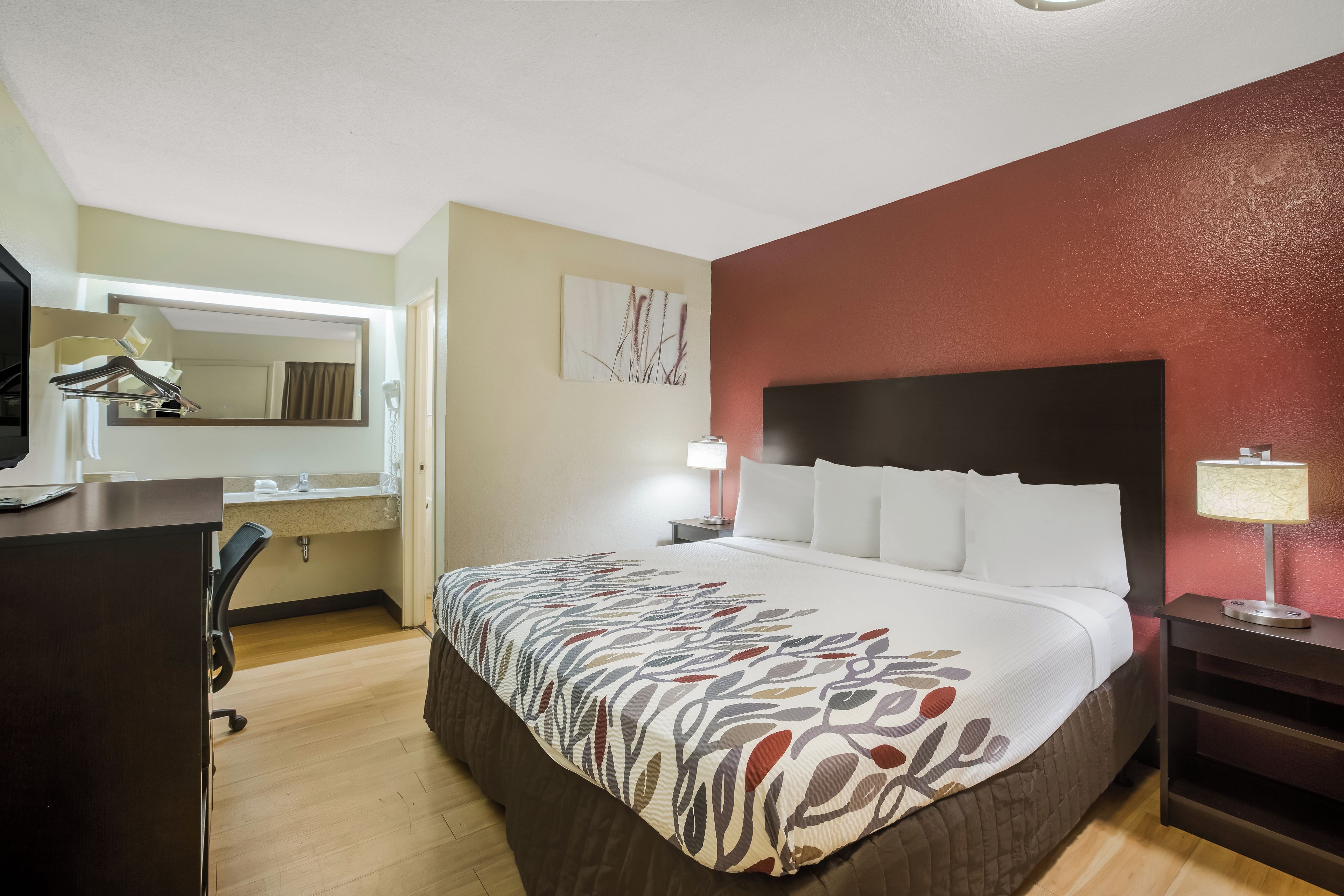A hotel room with a bed, a desk with a chair, a mirror, a nightstand with a lamp, and a wall with a painting.