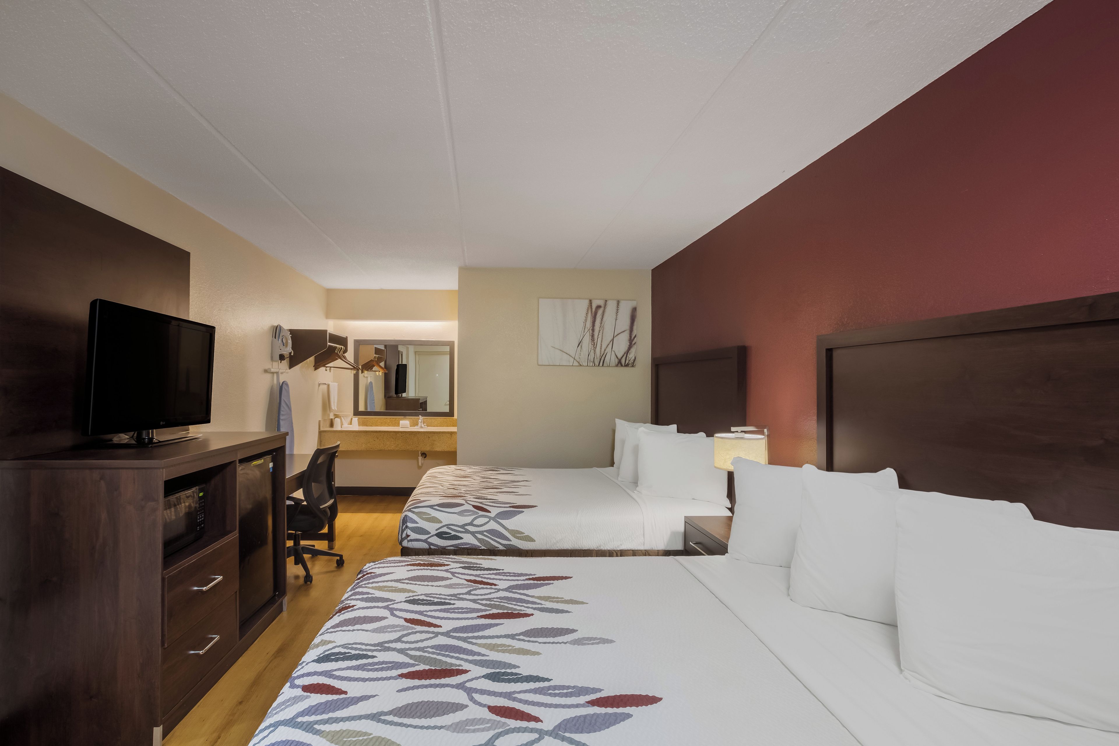 A hotel room with a bed, a desk with a television, a mirror, and a bathroom visible in the background.