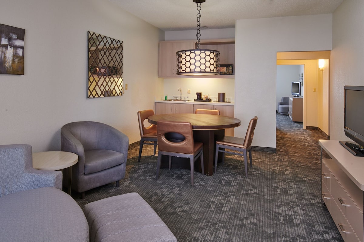 A hotel room with a dining area, a sofa, a TV, and a carpeted floor.