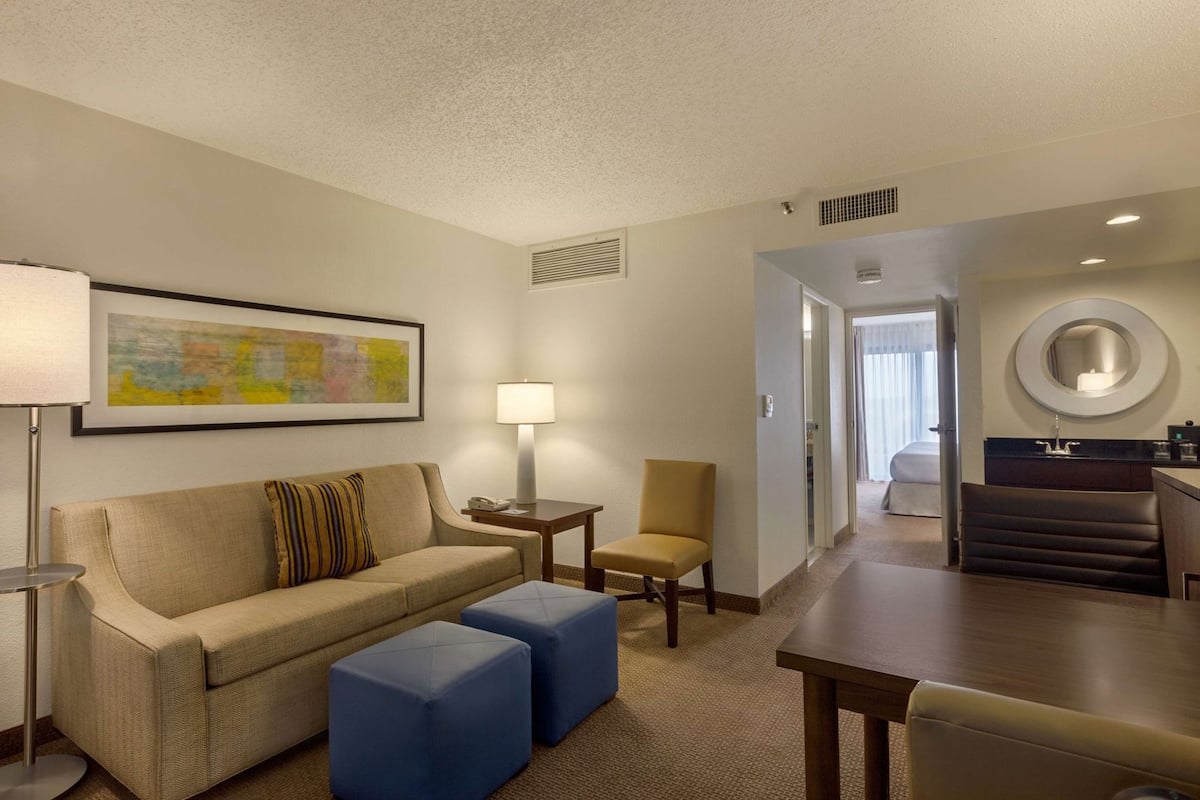 A hotel room with a sofa, ottomans, a lamp, a chair, a dining table, and a kitchen area.