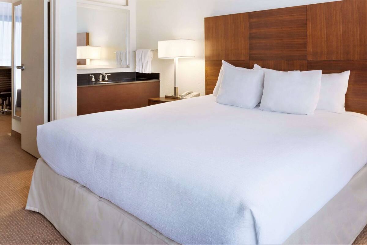 A hotel room with a large bed, a wooden headboard, a bedside table with a lamp, and a bathroom visible through an open door.
