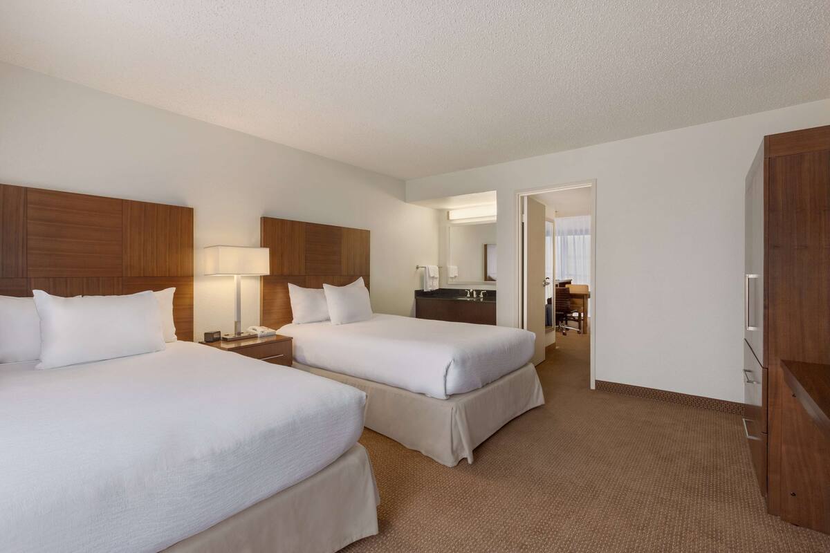 A hotel room with two beds, a desk with a lamp, a mirror, and a bathroom visible through an open door.