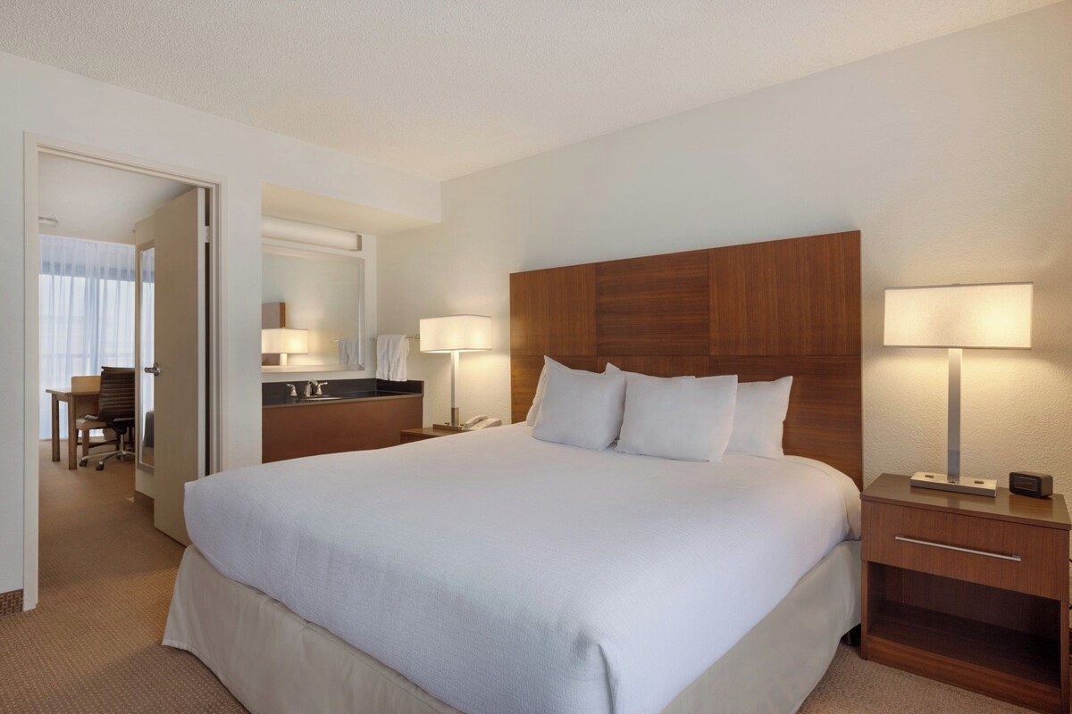 Une chambre d’hôtel avec un grand lit, des lampes de chevet, une tête de lit en bois, un bureau avec une chaise et une salle de bain équipée d’un miroir et d’un porte-serviettes.