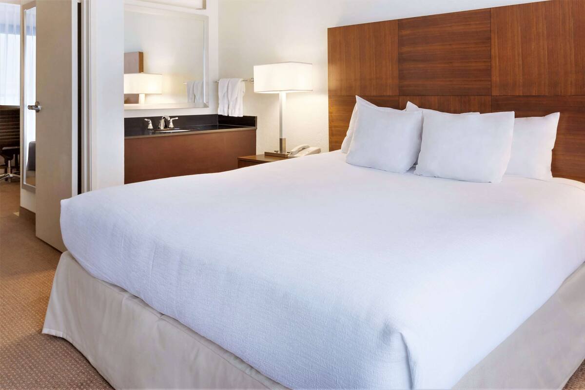 A hotel room with a large bed, a wooden headboard, a bedside table with a lamp, and a bathroom visible through an open door.