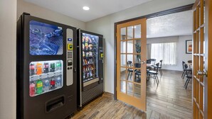 A room with a wooden floor, a vending machine with beverages, and a door leading to a dining area with chairs and tables. - Quality Inn Los Lunas - Albuquerque South (Los Lunas)