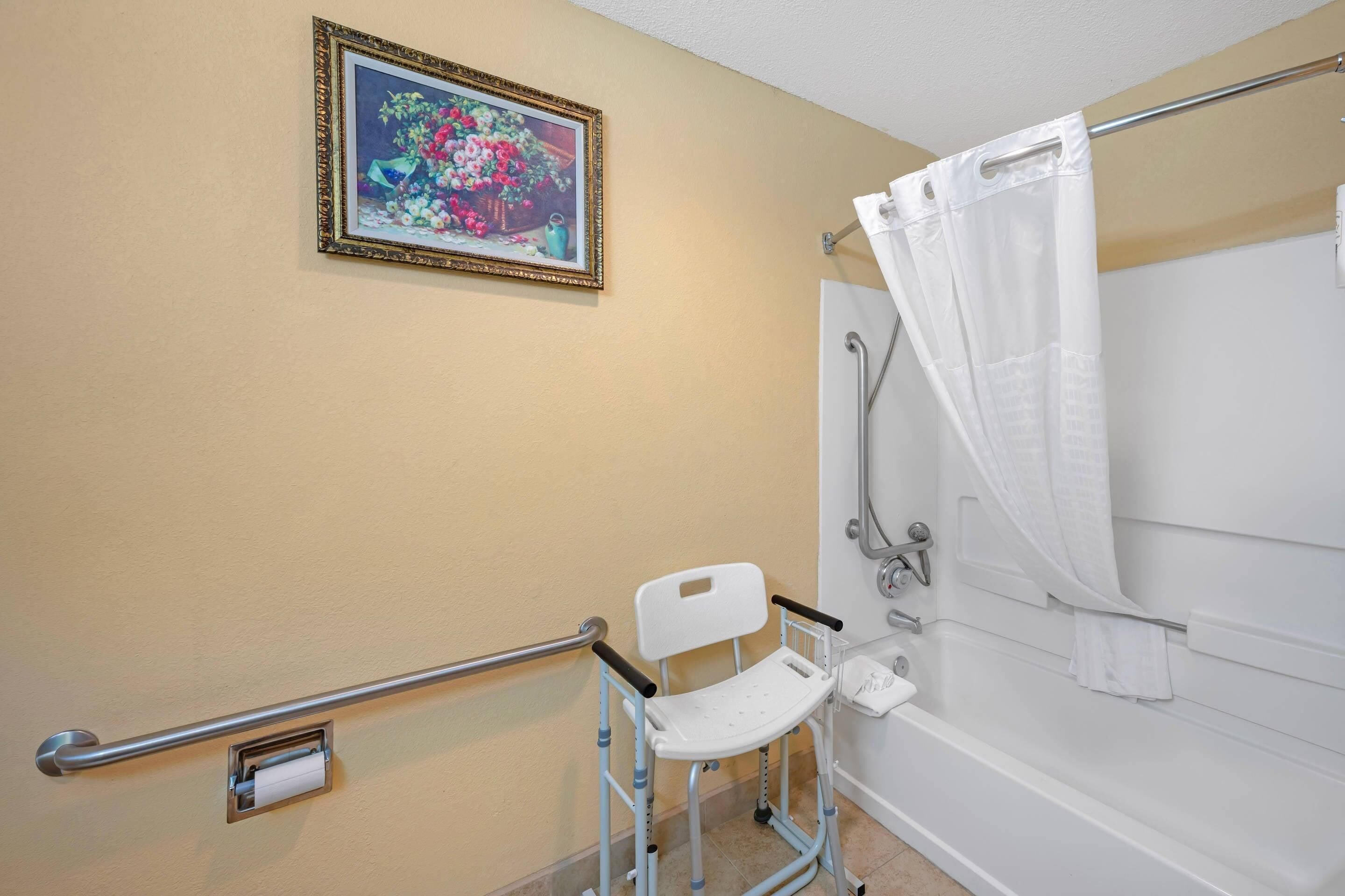 A bathroom with a shower, a white chair, a handrail, and a framed picture on the wall.