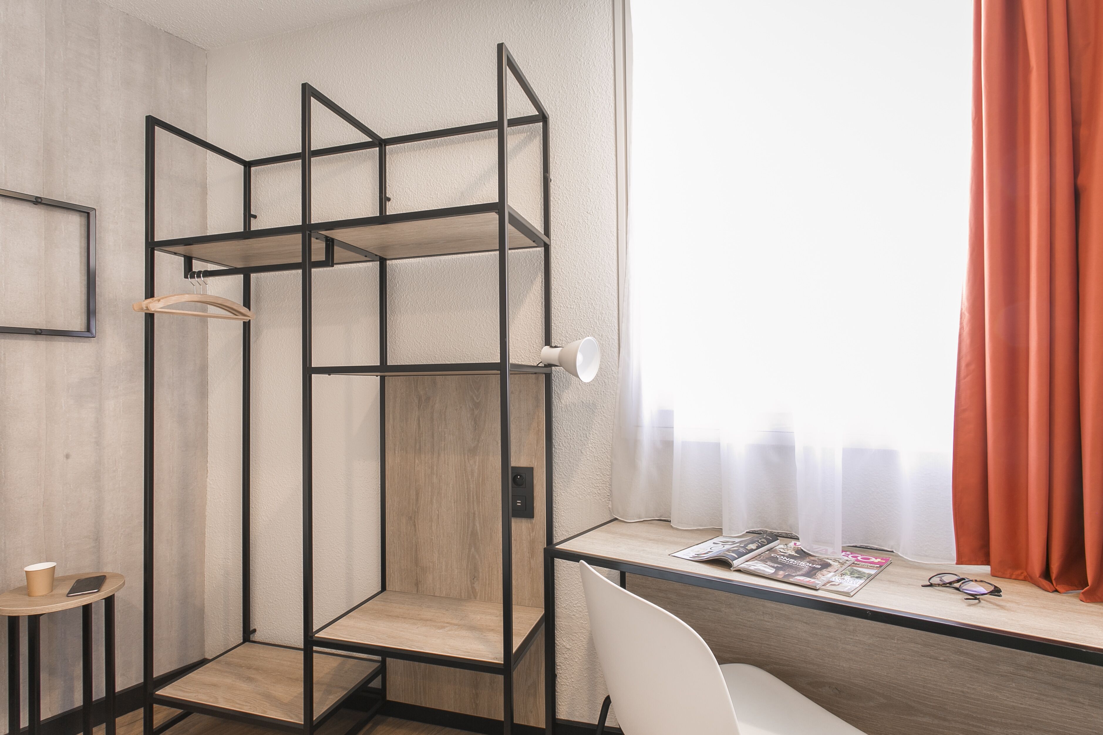 A modern room with a wooden desk, a white chair, a hanging coat rack, and a window with sheer curtains.