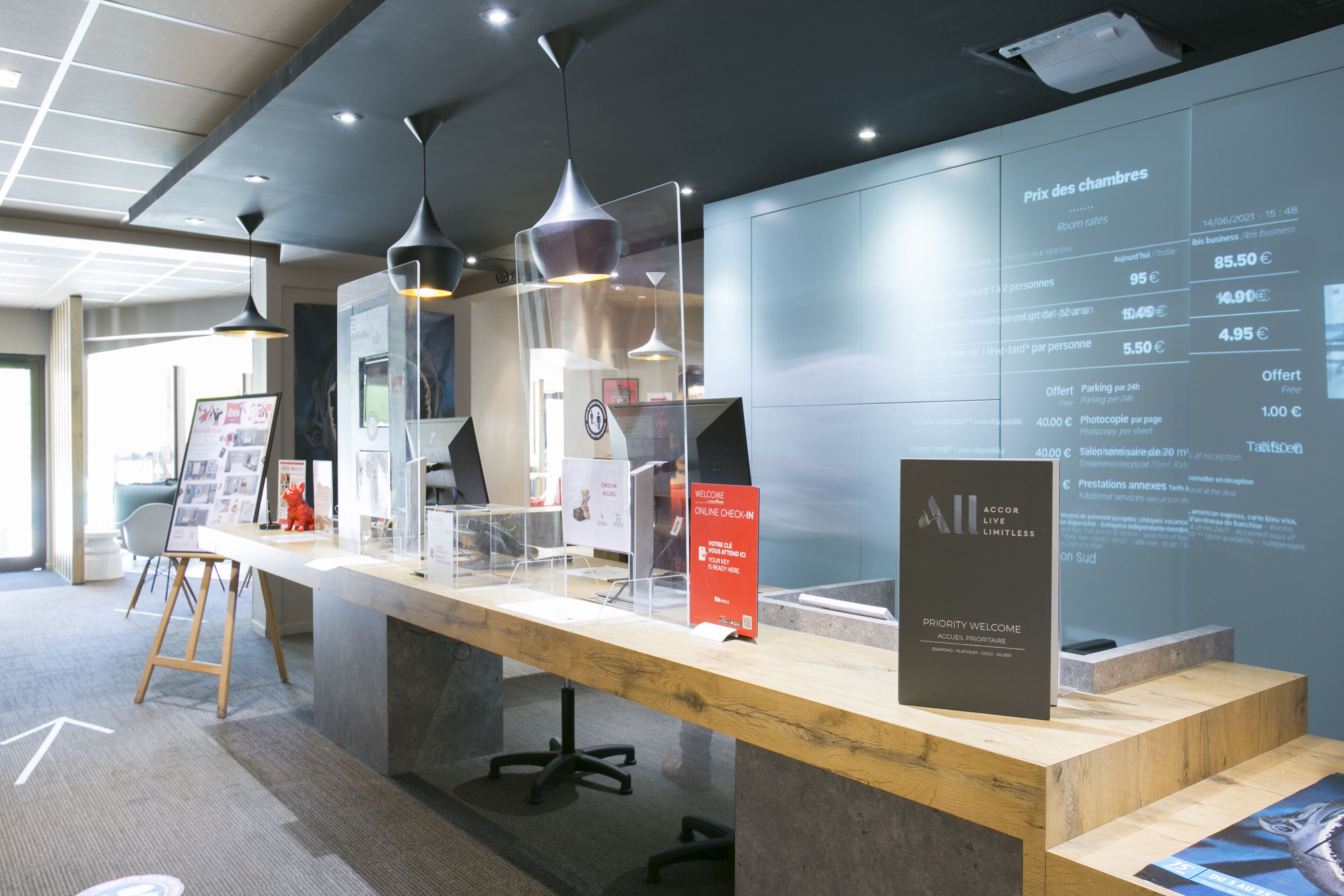 Un comptoir d’accueil avec un plan de travail en bois, des suspensions noires et un écran numérique affichant les tarifs des chambres.