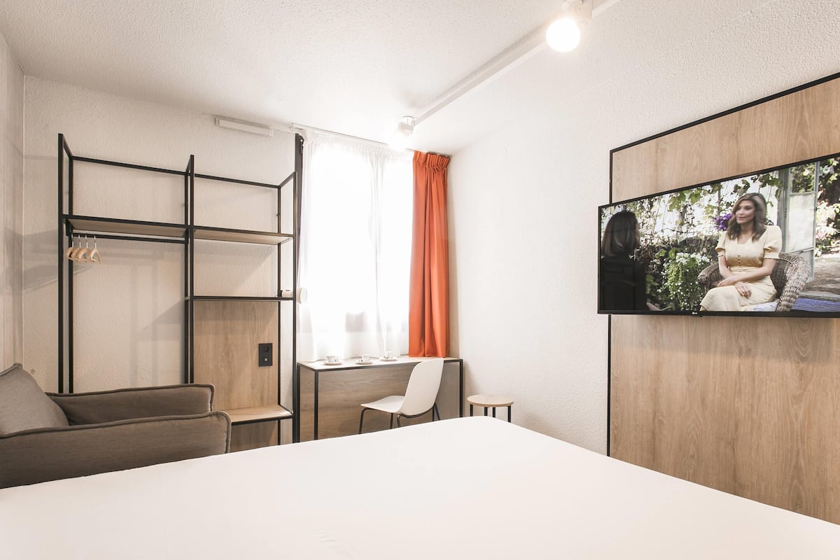 Une chambre d’hôtel moderne équipée d’un lit, d’un bureau, d’une chaise, d’une télévision et d’une fenêtre avec des rideaux.