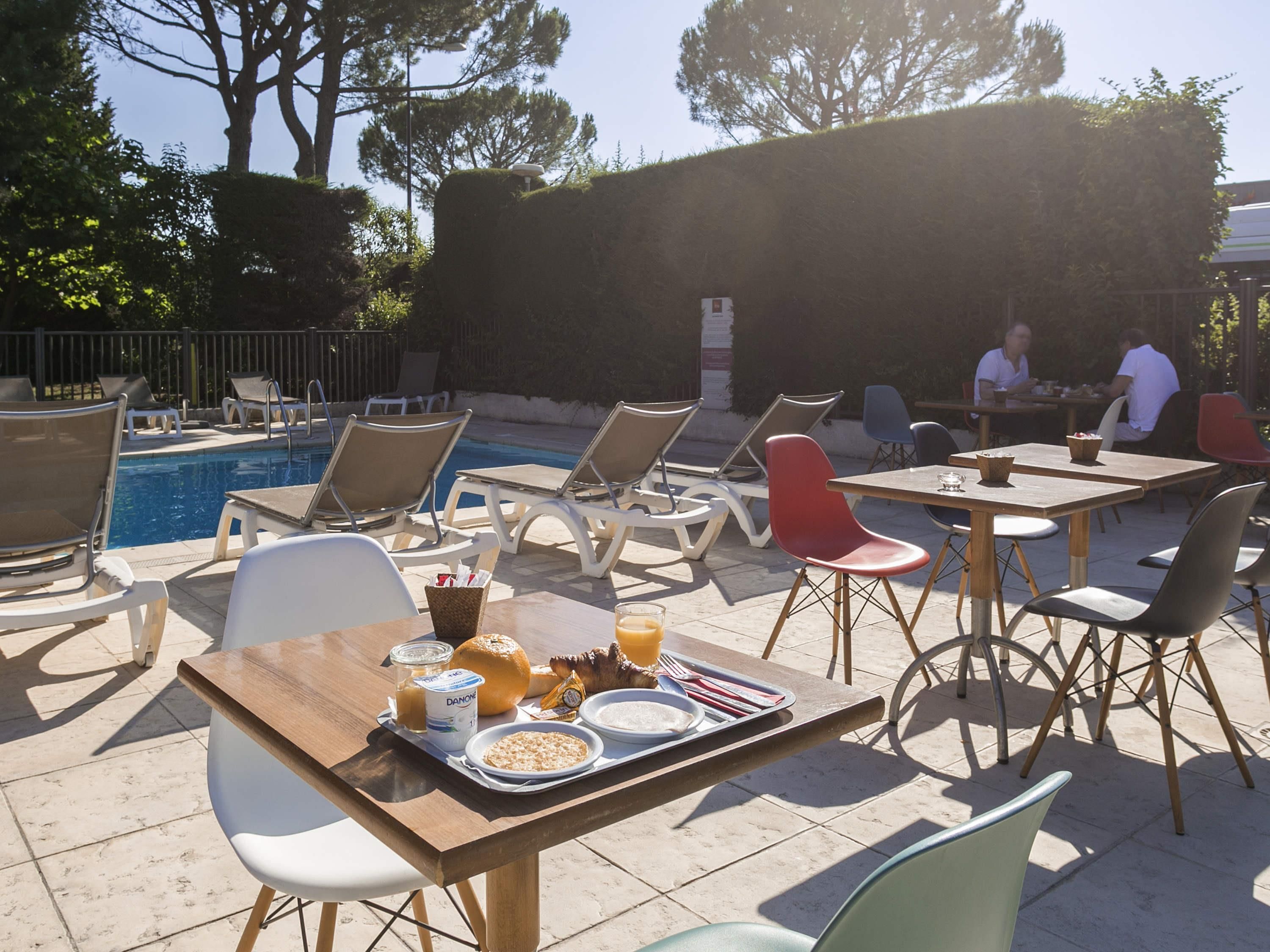 A poolside dining area with chairs and tables, a pool, and a hedge in the background.
