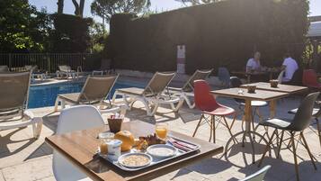 A poolside dining area with chairs and tables, a pool, and a hedge in the background.