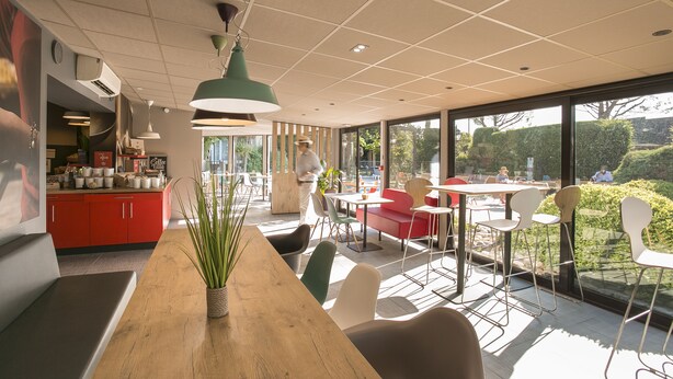 A modern cafe with a wooden table, red and white chairs, and a large window offering a view of the outdoors. - ibis Avignon Sud (Avignon)
