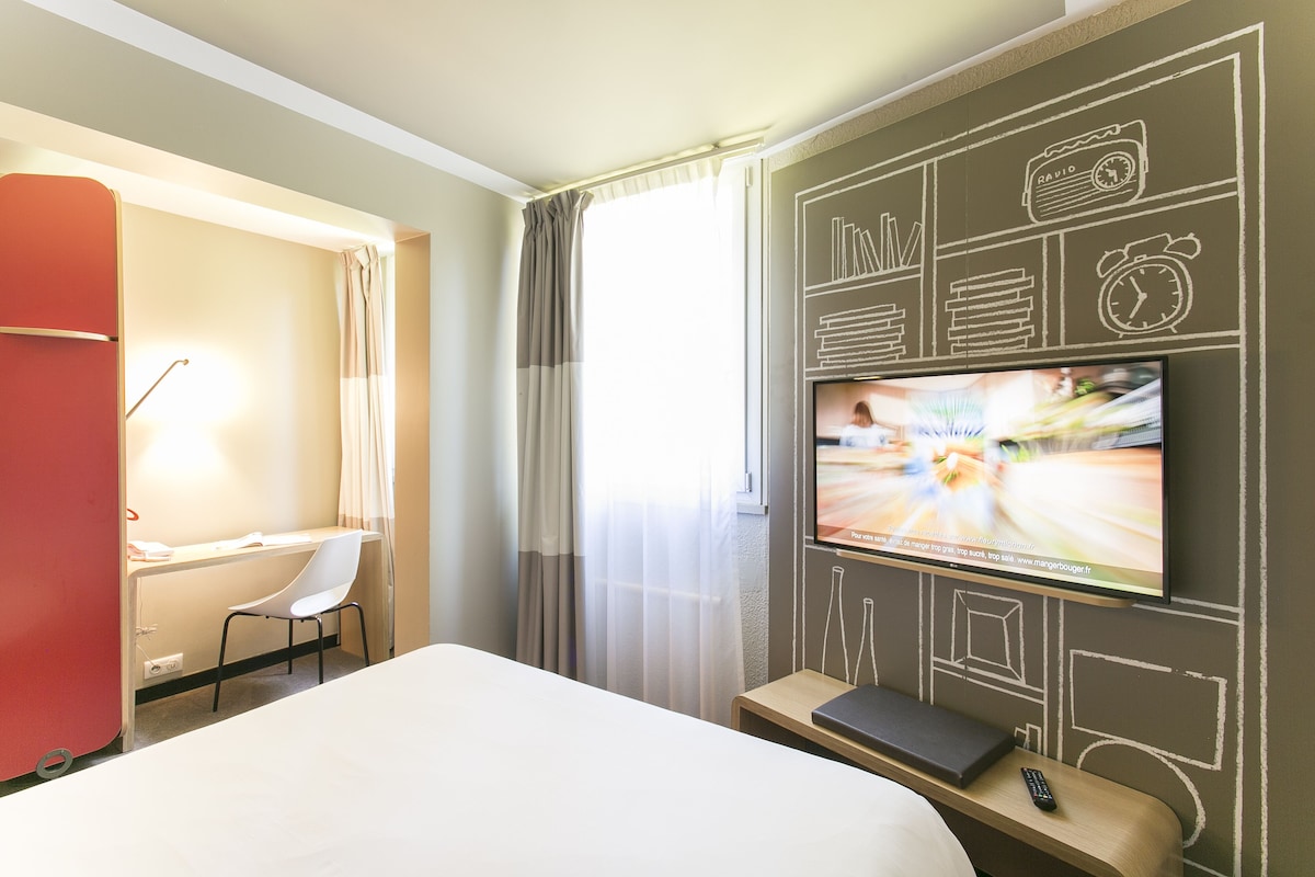 A hotel room with a bed, a desk with a chair, a TV mounted on the wall, and a window with curtains.