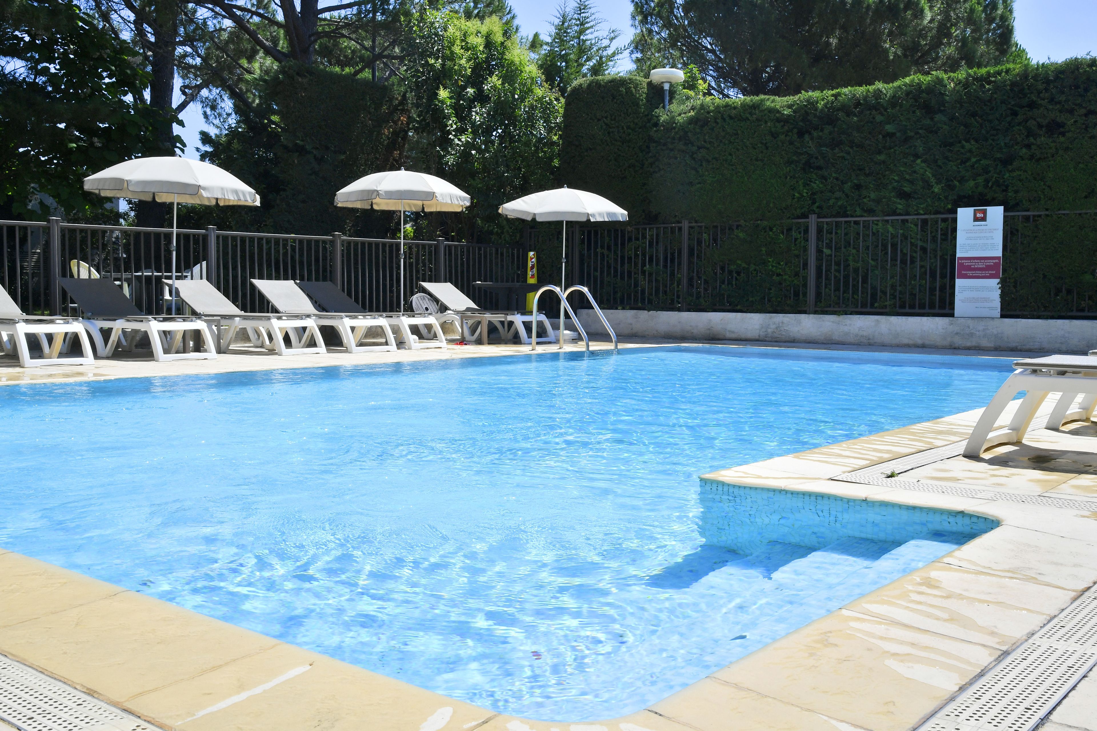 A swimming pool with lounge chairs and umbrellas, surrounded by a fence and trees.