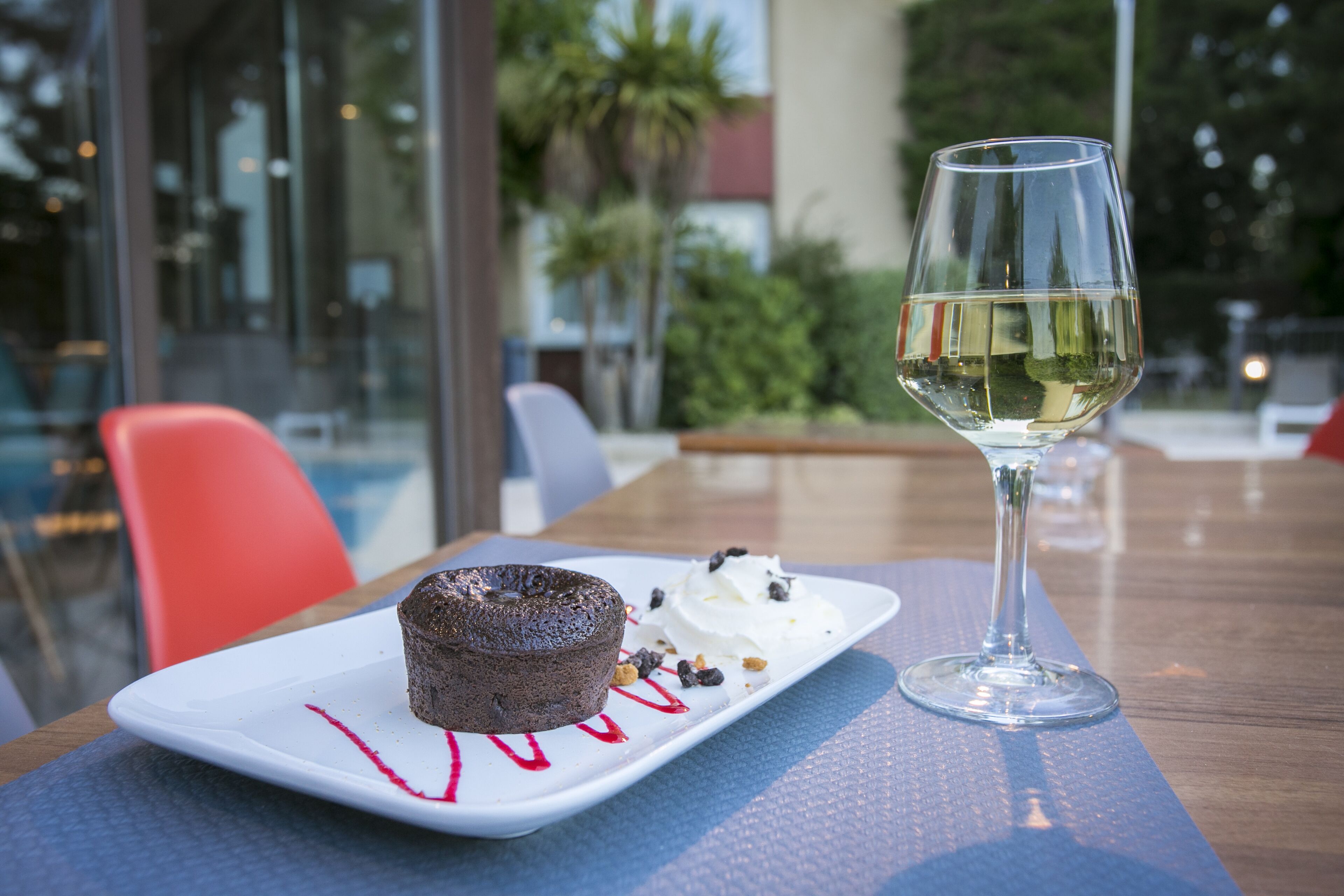 En desserttallrik med en chokladkaka och vispad grädde, tillsammans med ett glas vitt vin, står på ett bord utomhus.