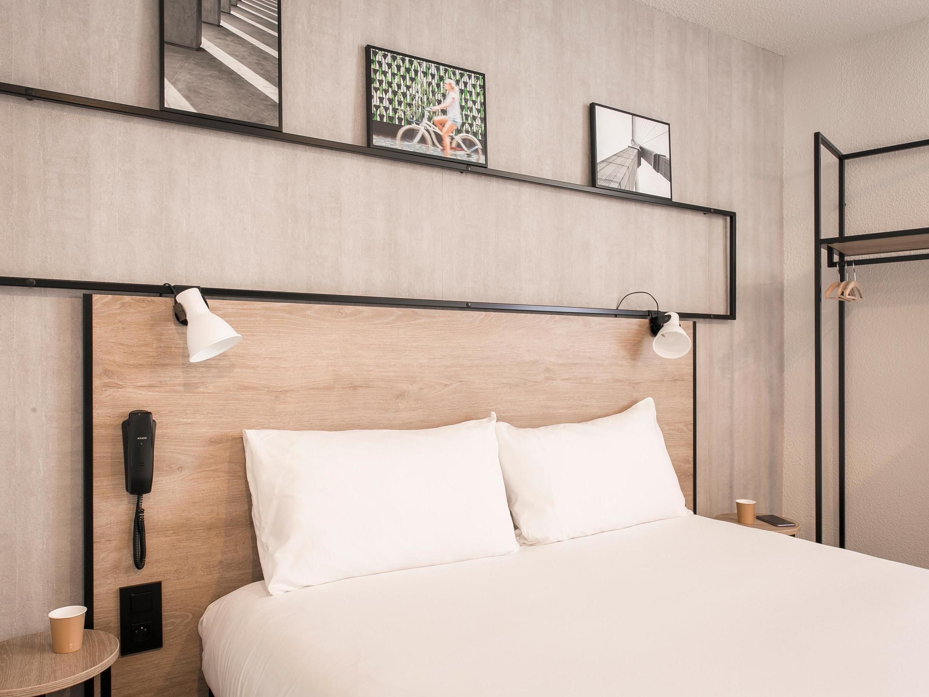 Habitación de hotel con cabecera de madera, cama con ropa blanca, teléfono en la mesita de noche, una lámpara y una pequeña mesa con una taza.