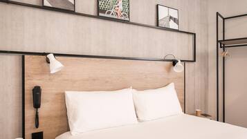 Habitación de hotel con cabecera de madera, cama con ropa blanca, teléfono en la mesita de noche, una lámpara y una pequeña mesa con una taza.