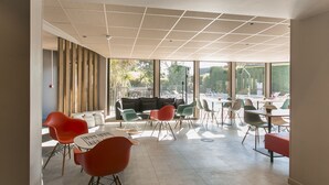 A modern indoor space with a mix of seating options including sofas and chairs, large windows providing natural light, and a view of the outdoors. - ibis Avignon Sud (Avignon)