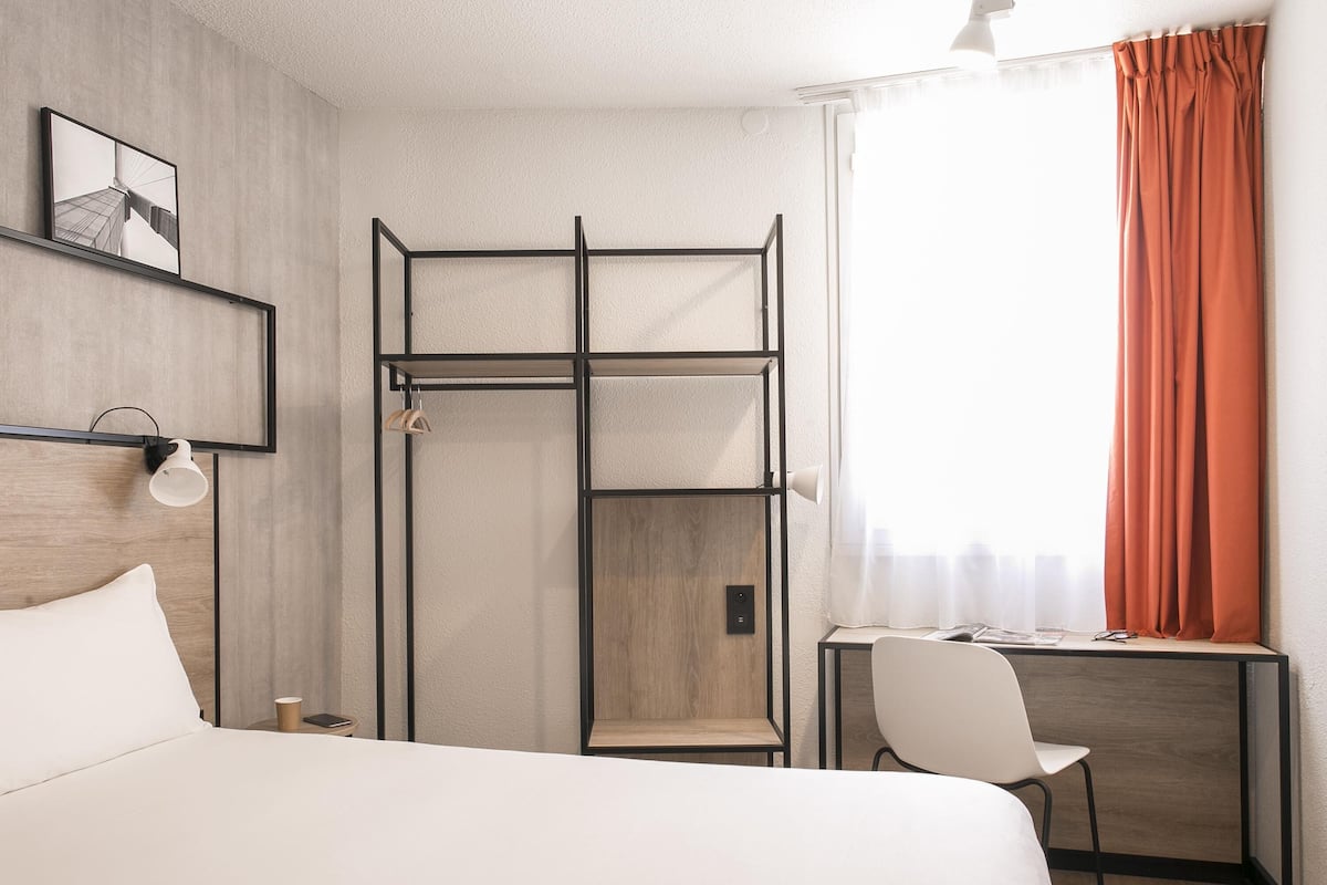 A hotel room with a bed, a desk with a chair, a shelf unit, and a window with curtains.