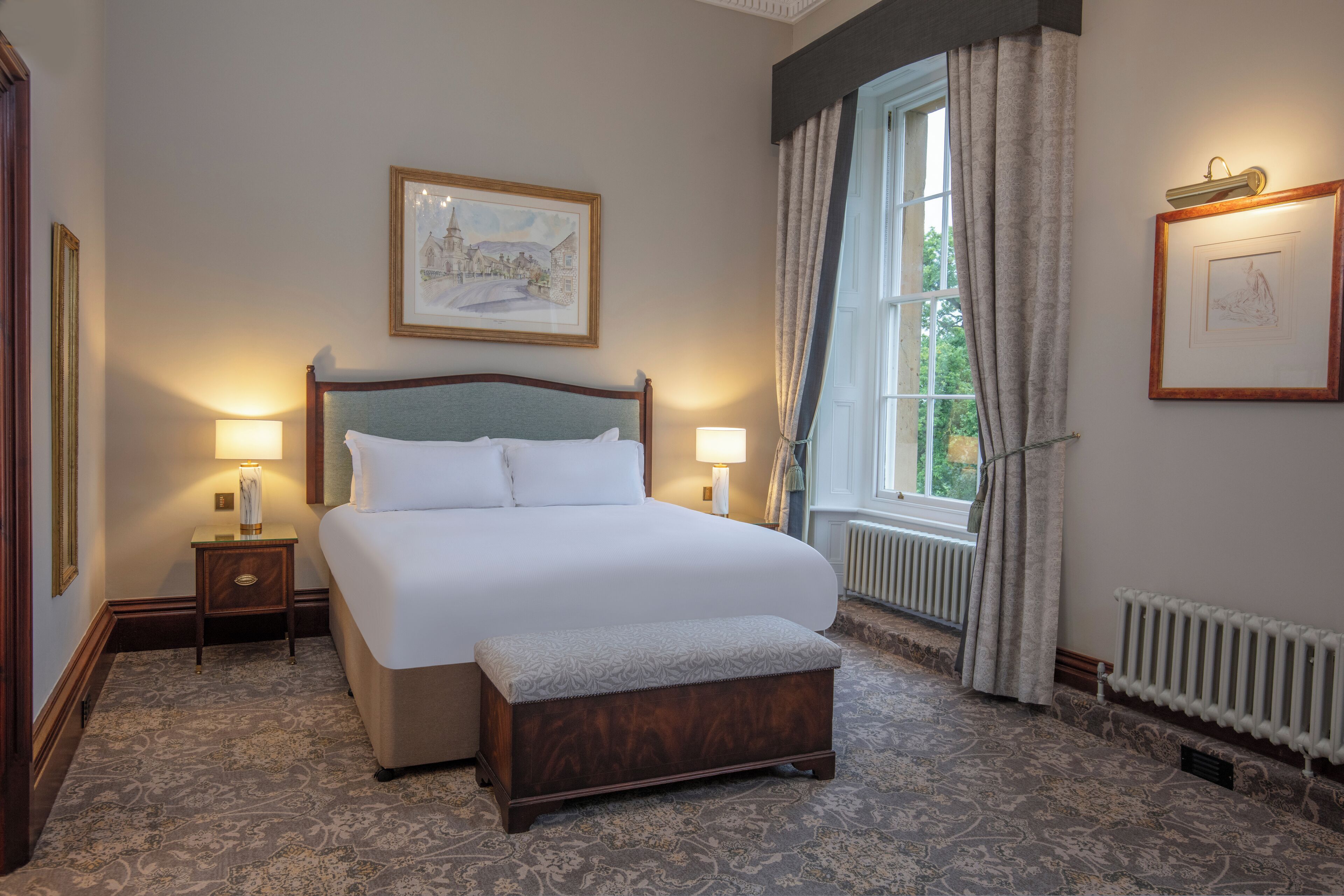 A neatly arranged hotel room with a large bed, two bedside lamps, a framed picture on the wall, a window with curtains, and a radiator below the window.
